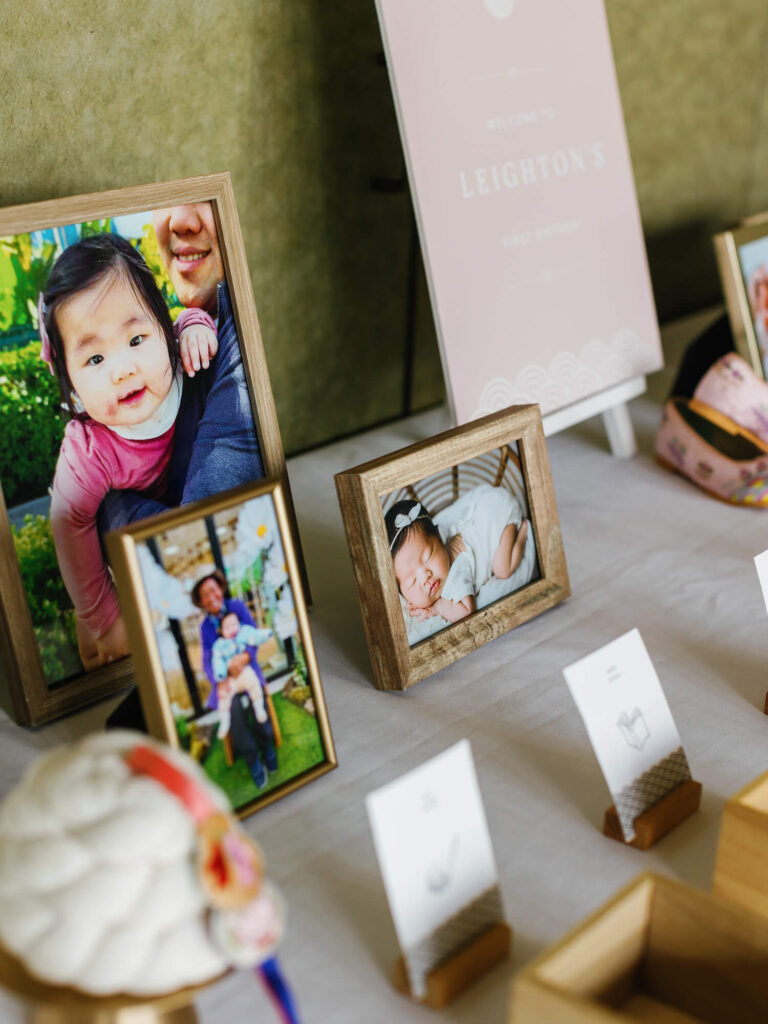 Framed photos of a baby and family are displayed on a table. A sign reads "Leighton's First Birthday." The scene is joyful and celebratory.