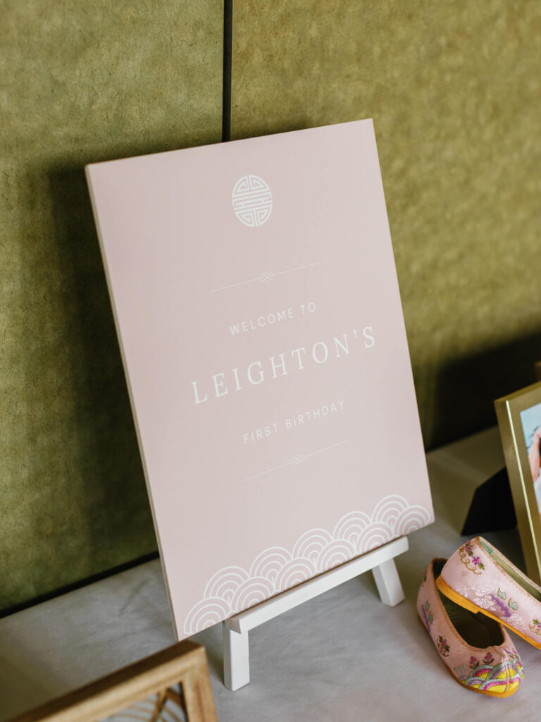 A pastel pink sign on an easel reads "Welcome to Leighton's First Birthday" in white text, with decorative elements. Nearby, there are floral-patterned shoes.