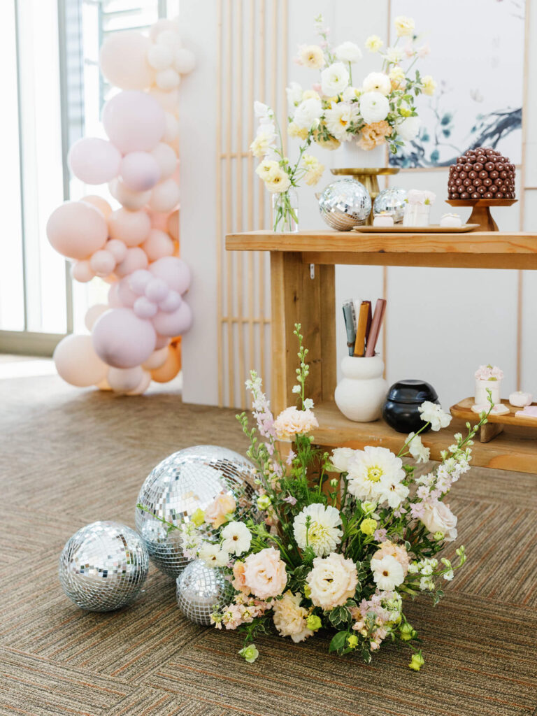 A wooden table adorned with a floral arrangement and disco balls sits beside pastel balloons. The setup conveys a festive and elegant atmosphere.