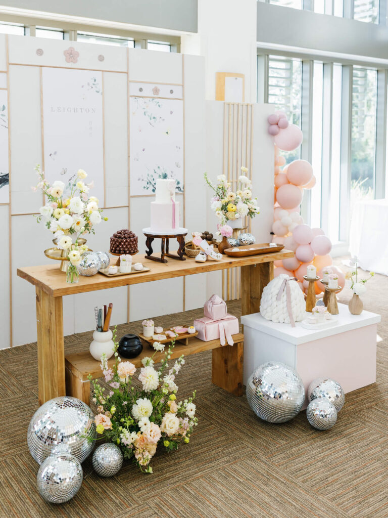 Elegant dessert table with flowers, candles, and a cake, accented by silver disco balls and pink balloons. The setting is bright and festive.
