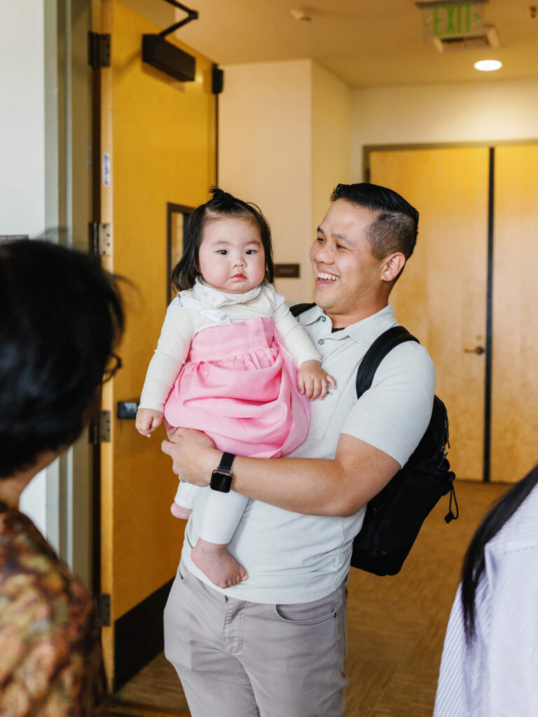 A smiling man holds a baby in a pink dress inside a room with wooden doors. The scene conveys warmth and joy, as he looks at someone off-camera.