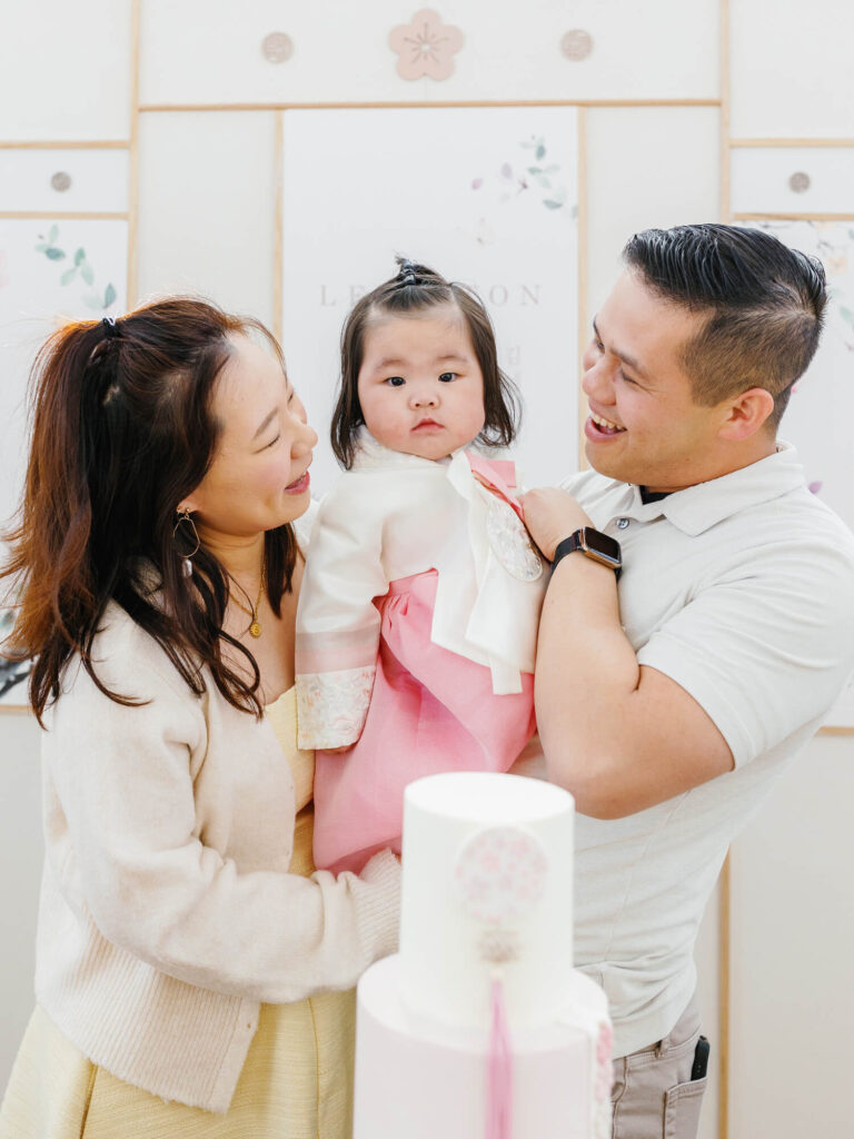 A family celebrating a special occasion with a baby in traditional attire. The parents are smiling joyfully at their child, with a decorative cake in front of them.