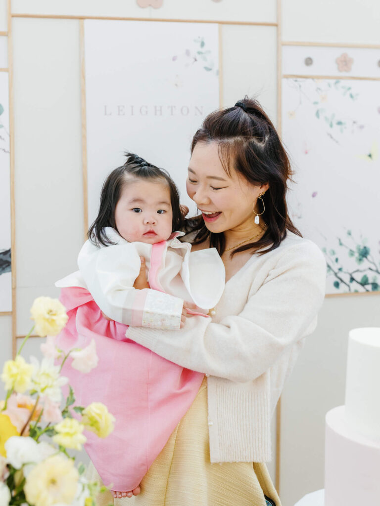 A woman joyfully holds a baby dressed in pink and white traditional attire against a soft floral backdrop. Yellow flowers in the foreground add warmth.