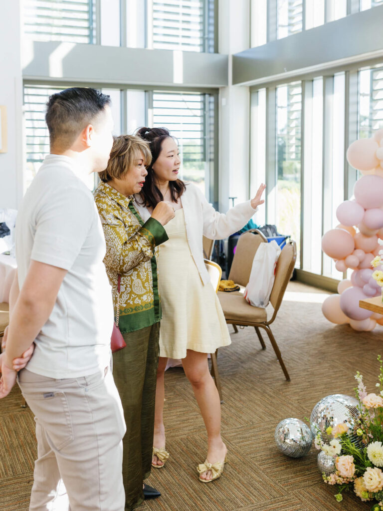 A woman in a light dress gestures while talking to another woman in green. A man stands nearby. They are in a sunlit room with balloons and disco balls.