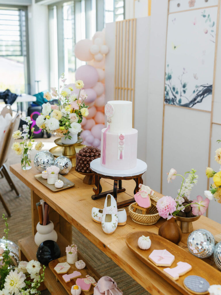 Elegant dessert table with a two-tiered white cake, surrounded by pastel flowers, small pastries, and decorative balloons.