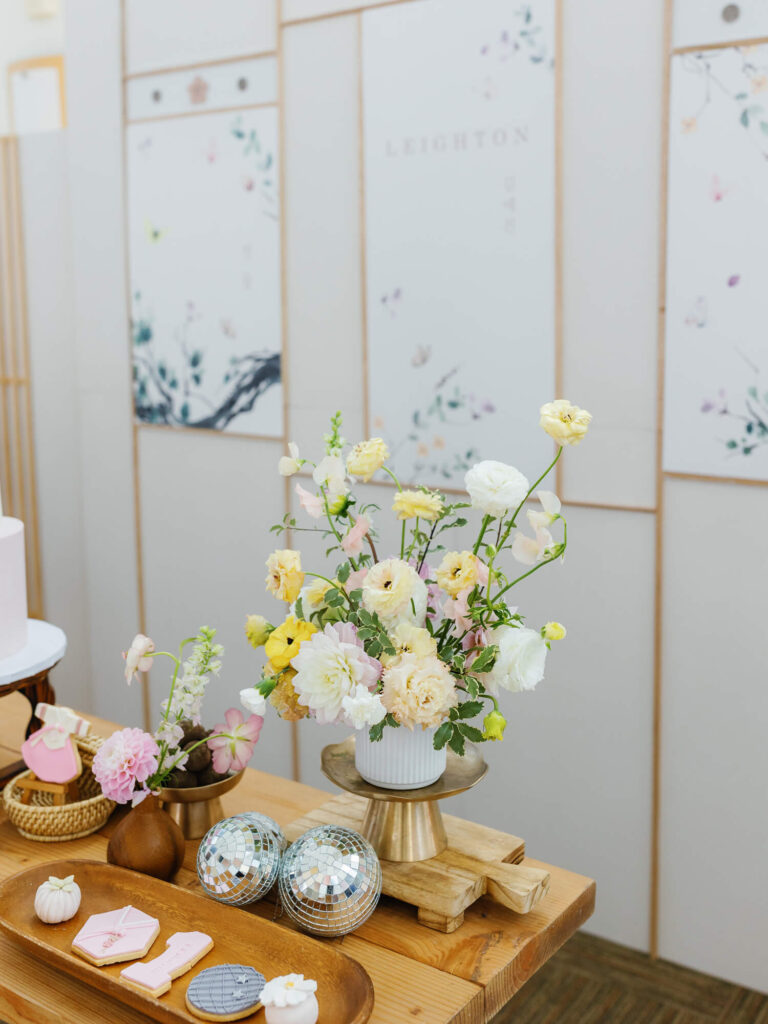 Elegant setting with pastel floral arrangement on a wooden table. Nearby, treats and disco balls add charm. Background features delicate floral panels.