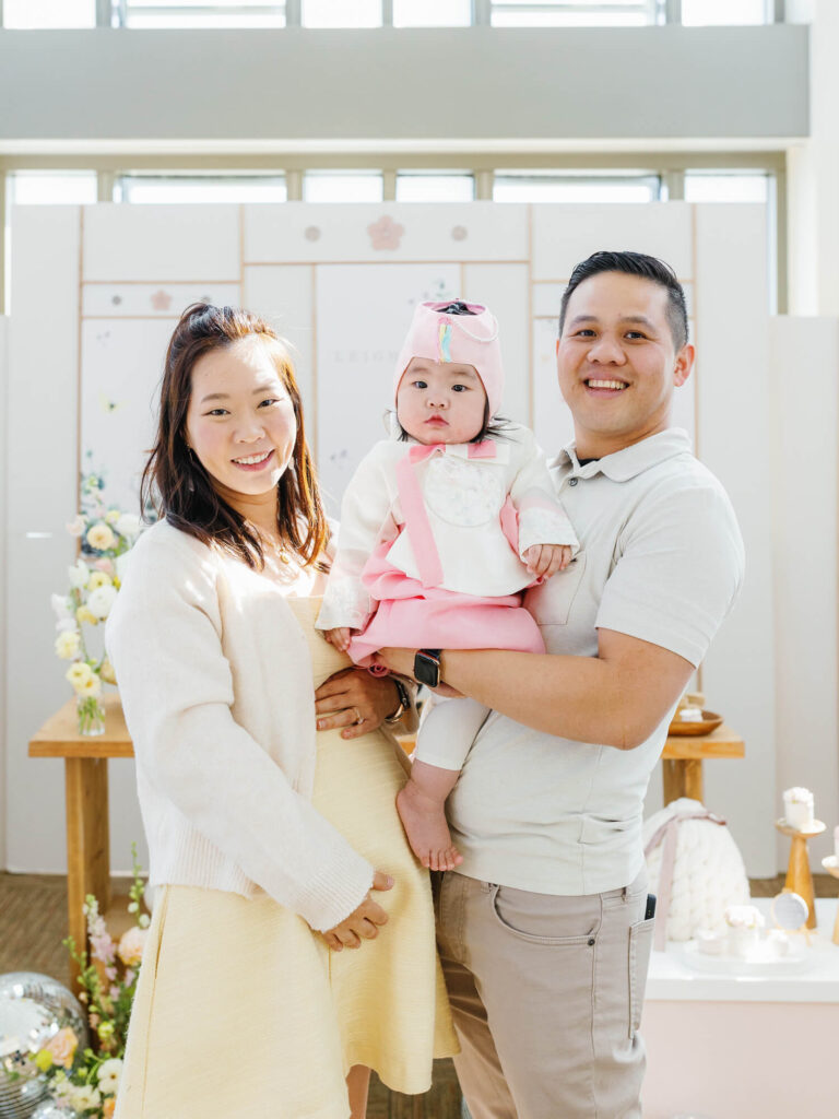 A joyful family photo at a celebration. A man and woman joyfully hold a baby in pink, standing behind a pink cake with yellow and white flowers around.
