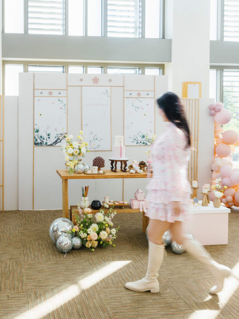 A woman in a pink dress and boots walks past a decorative table adorned with flowers, cakes, and balloons in a bright, modern room.