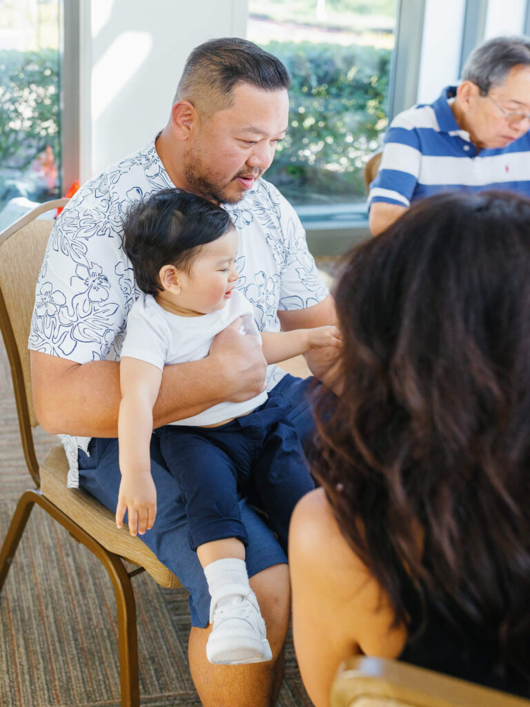 A man in a patterned shirt holds a smiling baby on his lap, while another person sits nearby. A man in a striped shirt is seated in the background.