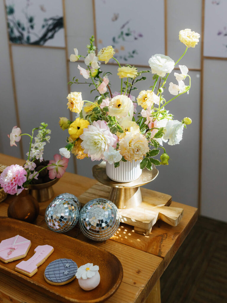 A vase with pastel yellow and pink flowers sits on a wooden table with decorative cookies and mini disco balls, creating a bright and cheerful scene.