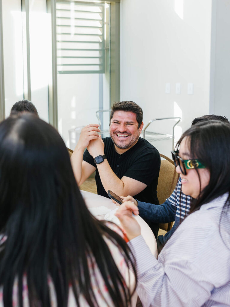 A group of people sit around a table in a brightly lit room. One person, smiling widely, engages in conversation.