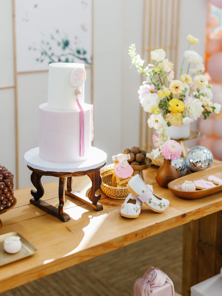 A two-tiered pastel cake on a wooden stand is surrounded by flowers, baby shoes, and assorted desserts on a table, creating a festive, elegant atmosphere.
