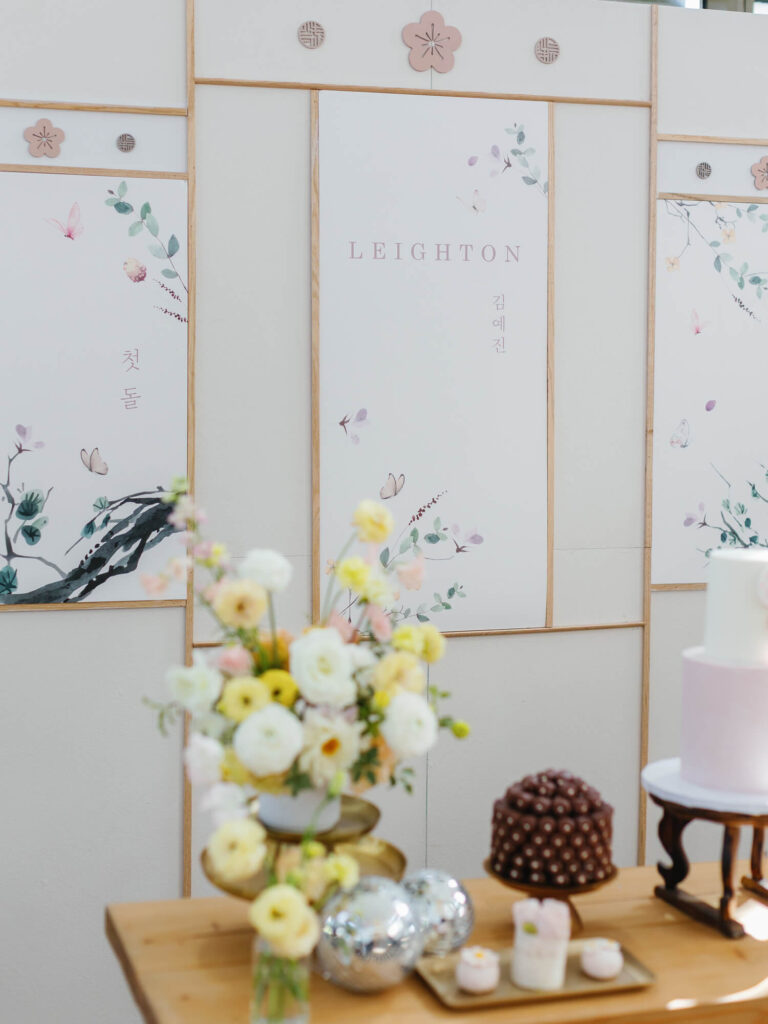 Decorative backdrop with floral artwork, butterfly motifs, and the word "LEIGHTON" in soft colors. A table in front holds a pastel flower arrangement, small cake, desserts, and mirrored spheres, creating an elegant and serene atmosphere.