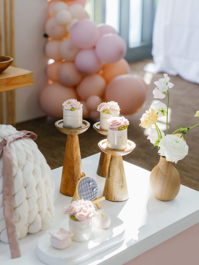Elegant dessert display featuring mini cakes with pink frosting on wooden pedestals, surrounded by soft pink balloons and a vase of white flowers.
