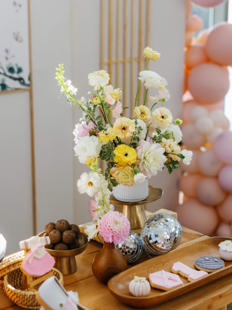Elegant table setting with a bouquet of pastel flowers, trays of cookies, a basket of chocolate balls, and disco balls.