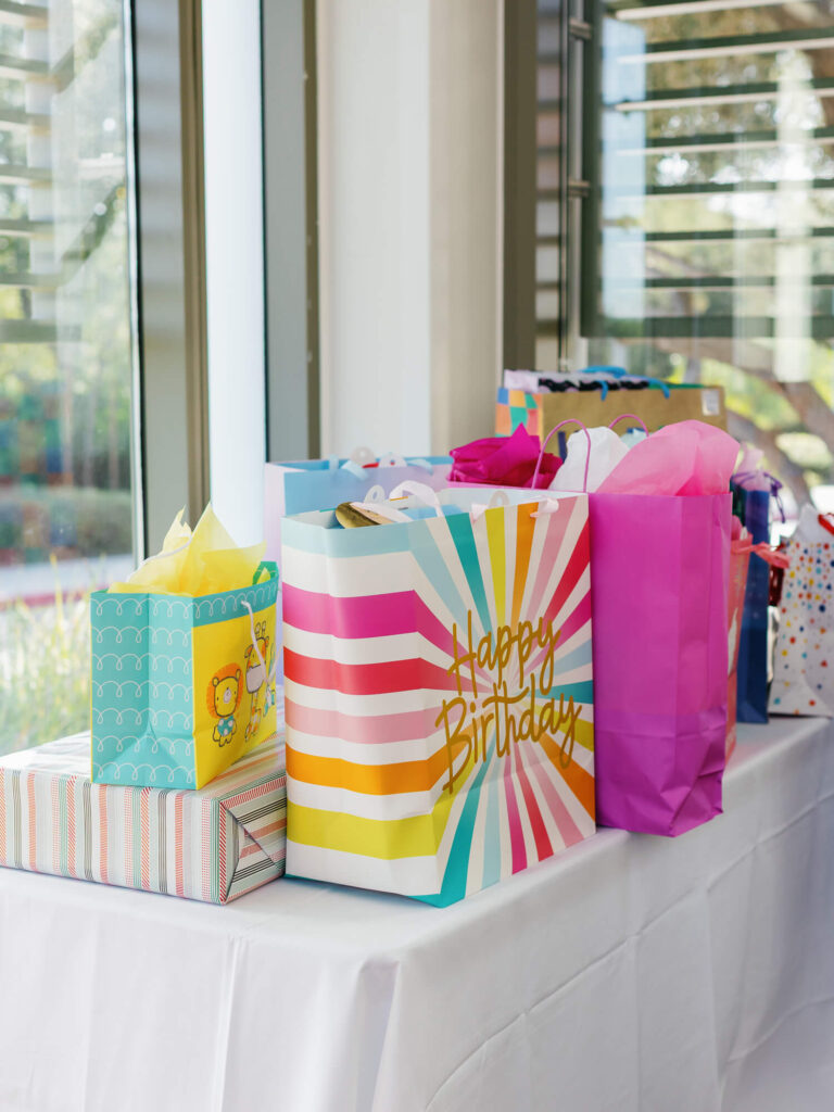 A collection of colorful birthday gift bags and boxes sits on a white tablecloth by a window.