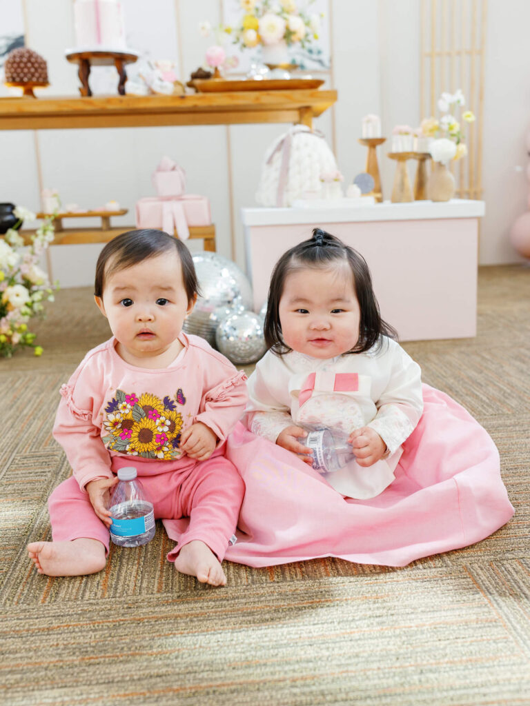 Two babies sitting on a carpet at a pink-themed party. The left baby wears a floral shirt, the right wears a pink dress, both holding water bottles.