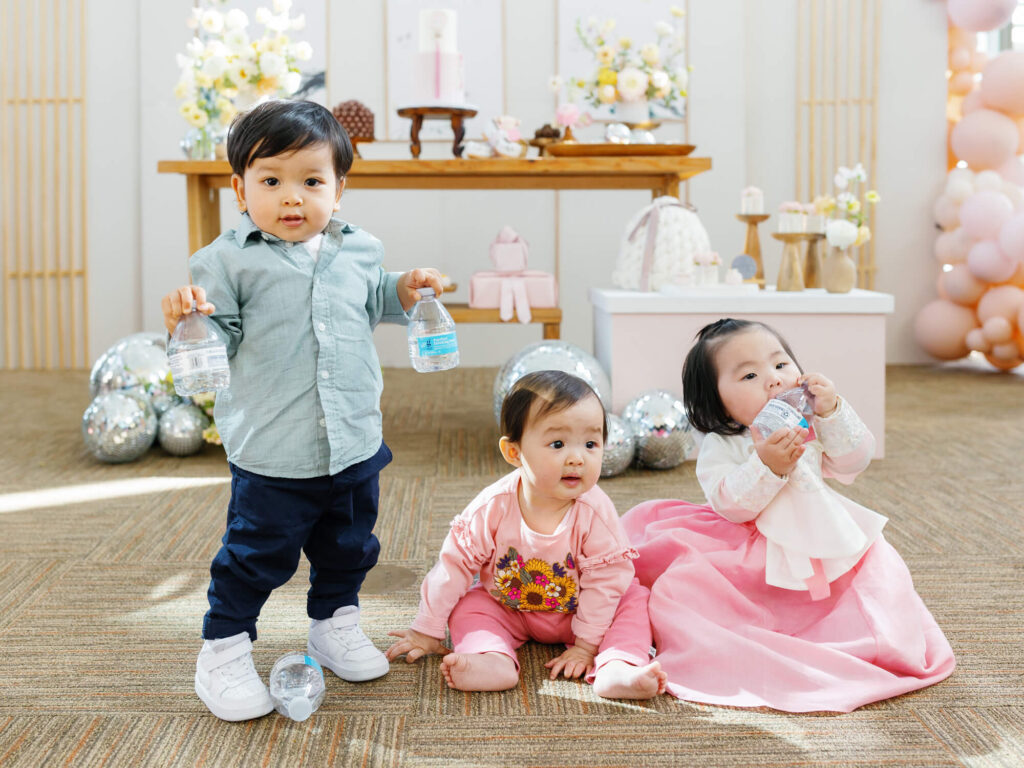 Three toddlers playfully engage at a party. One stands with water bottles, while the other two sit, one in a pink dress sipping water.