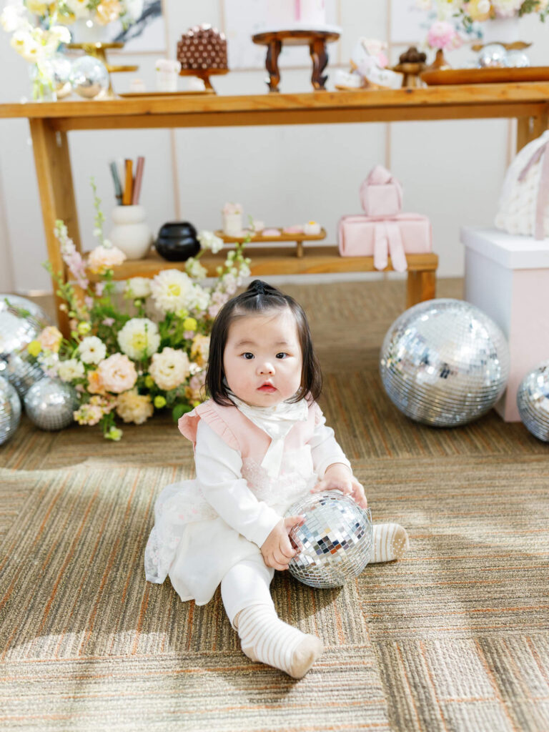 A young child in a white dress with pink accents sits on a carpet, holding a small disco ball. Behind, a wooden table features flowers and more disco balls, creating a festive atmosphere.