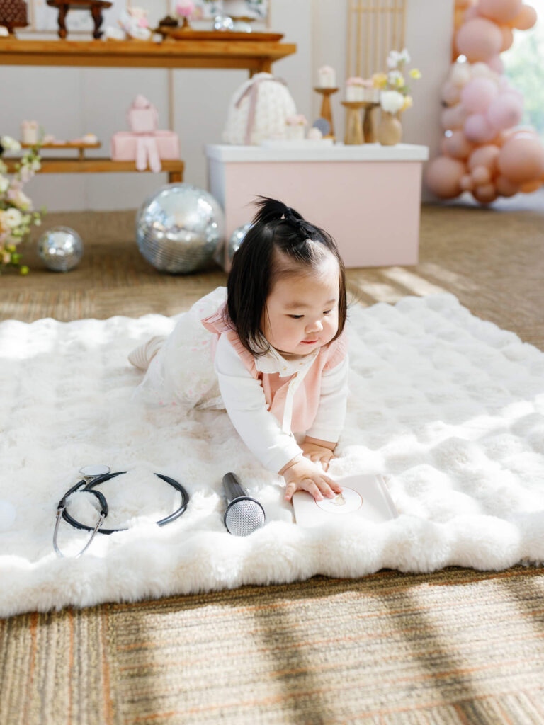 A baby, dressed in white and pink, crawls on a fluffy white rug, surrounded by a stethoscope, microphone, and writing tablet. Festive decor and balloons in the background.