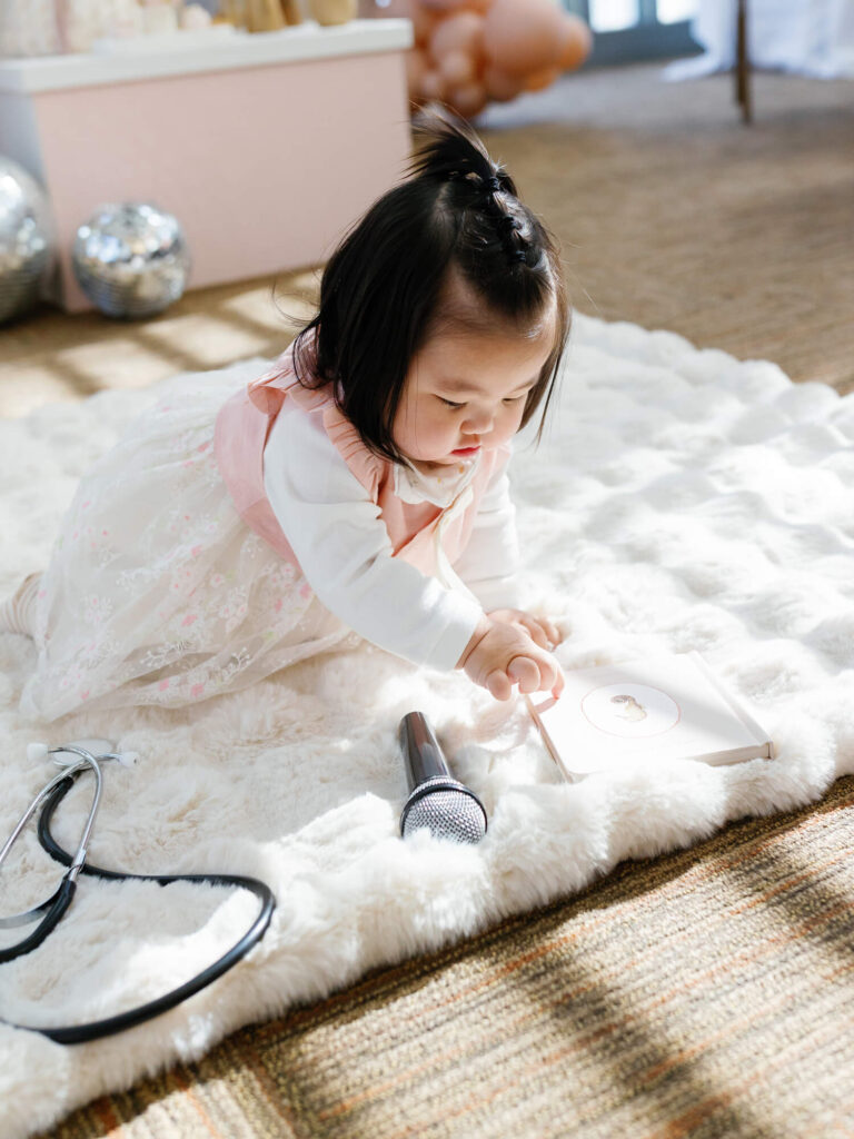A baby in a pink and white dress is crawling on a soft, white rug. Nearby are a microphone and stethoscope.