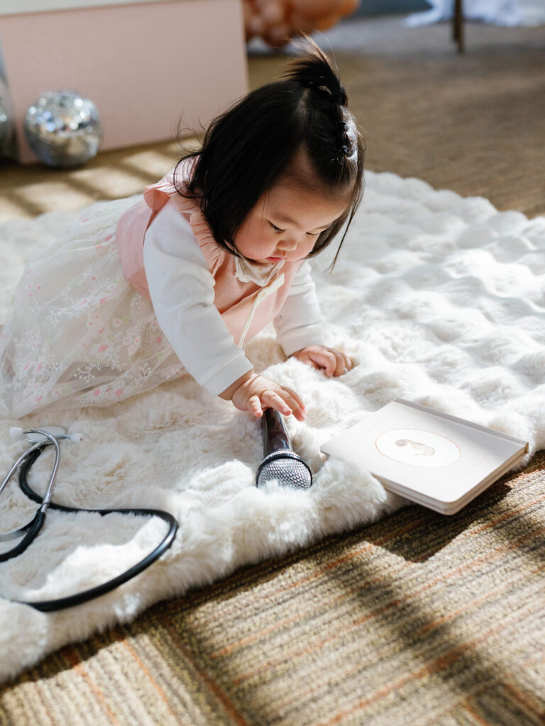 A young child in a white and pink dress plays on a soft rug, holding a microphone. Nearby are a book and a toy stethoscope.