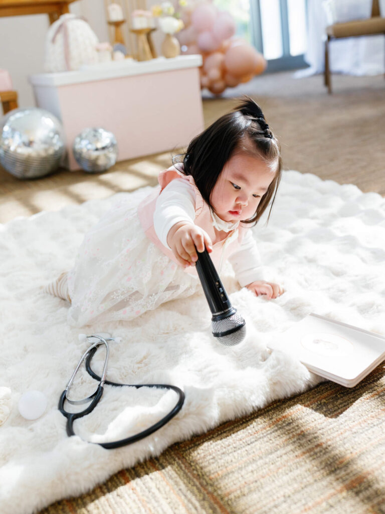 A toddler in a white dress lies on a fluffy rug, holding a microphone. Nearby are a stethoscope and pastel party decor, creating a playful scene.