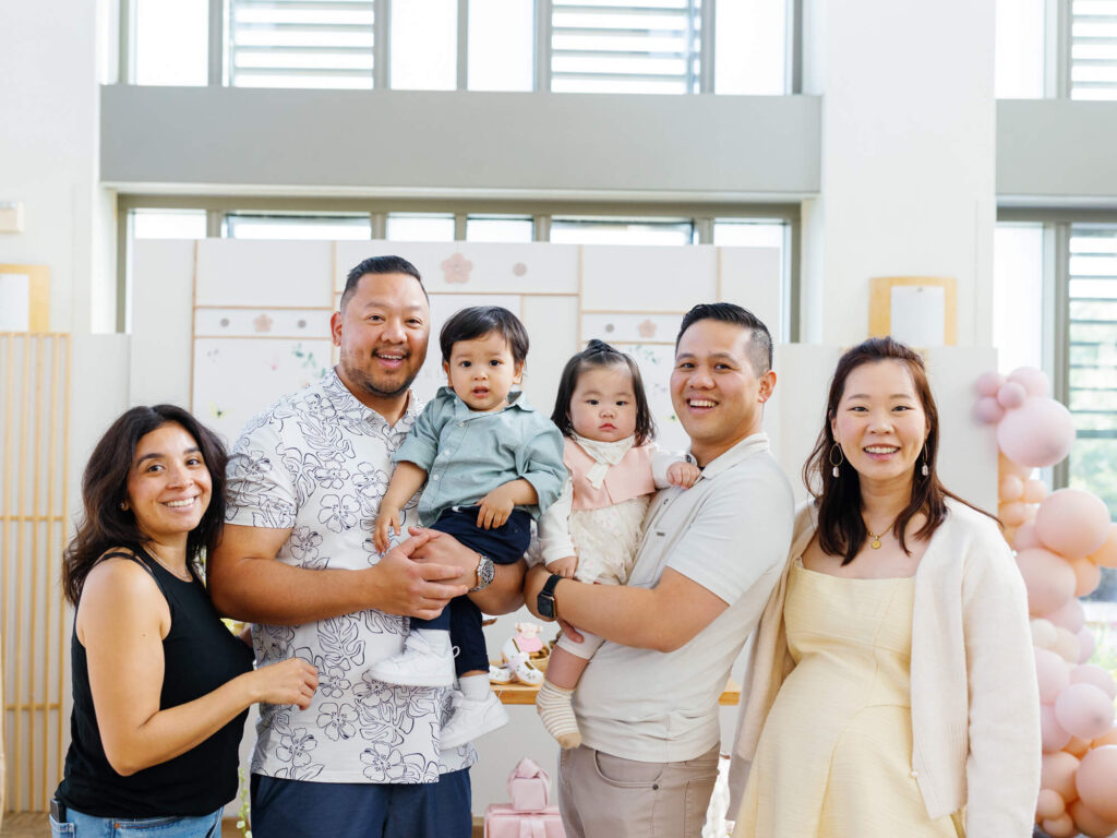 A cheerful group photo at a celebration. Five adults and two toddlers smile in a decorated room with pastel balloons and a sunny ambiance.